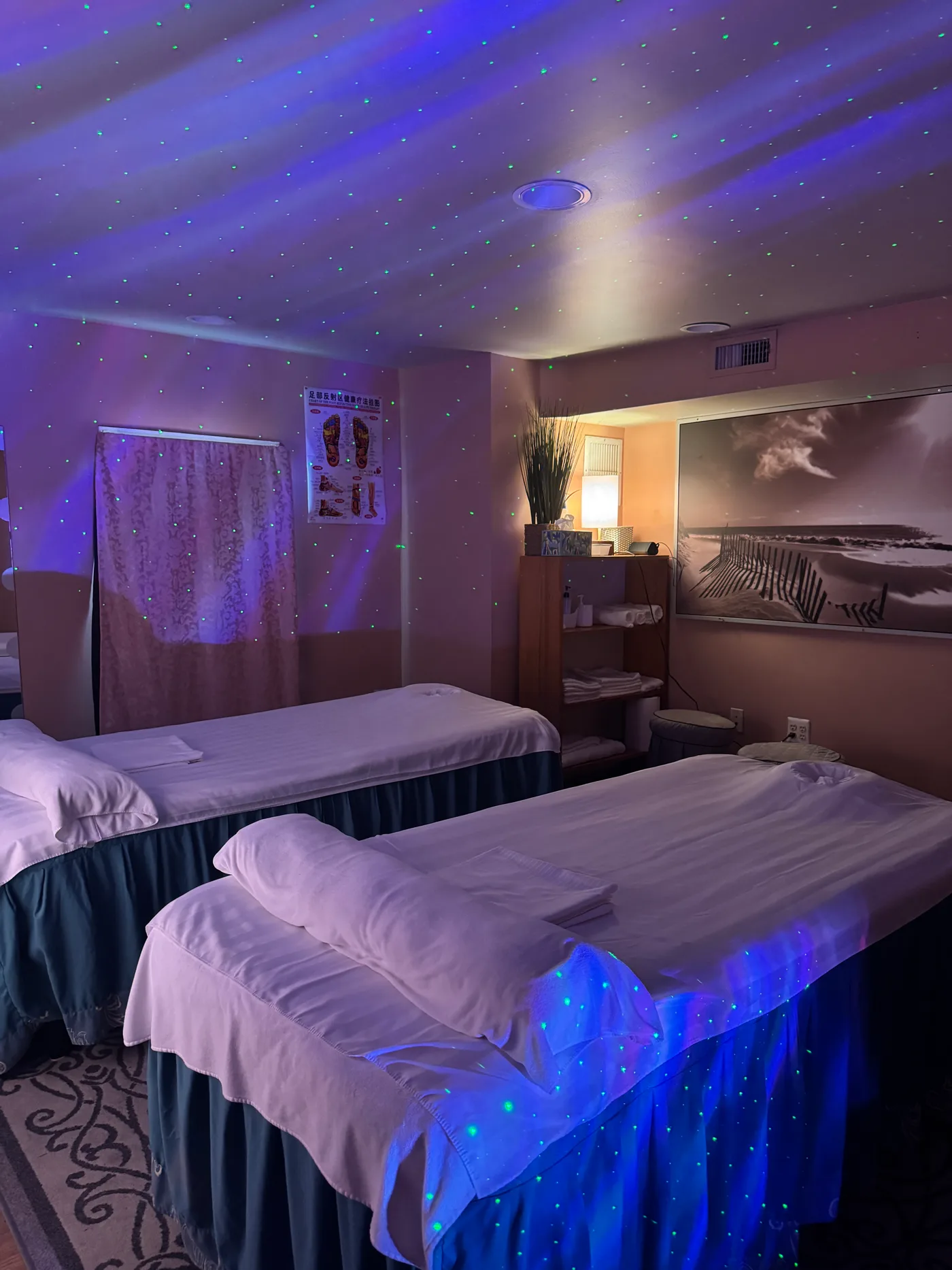 Two massage tables with white linens, teal skirts and a beach wall print at Wellness Foot Spa in Glenside, PA